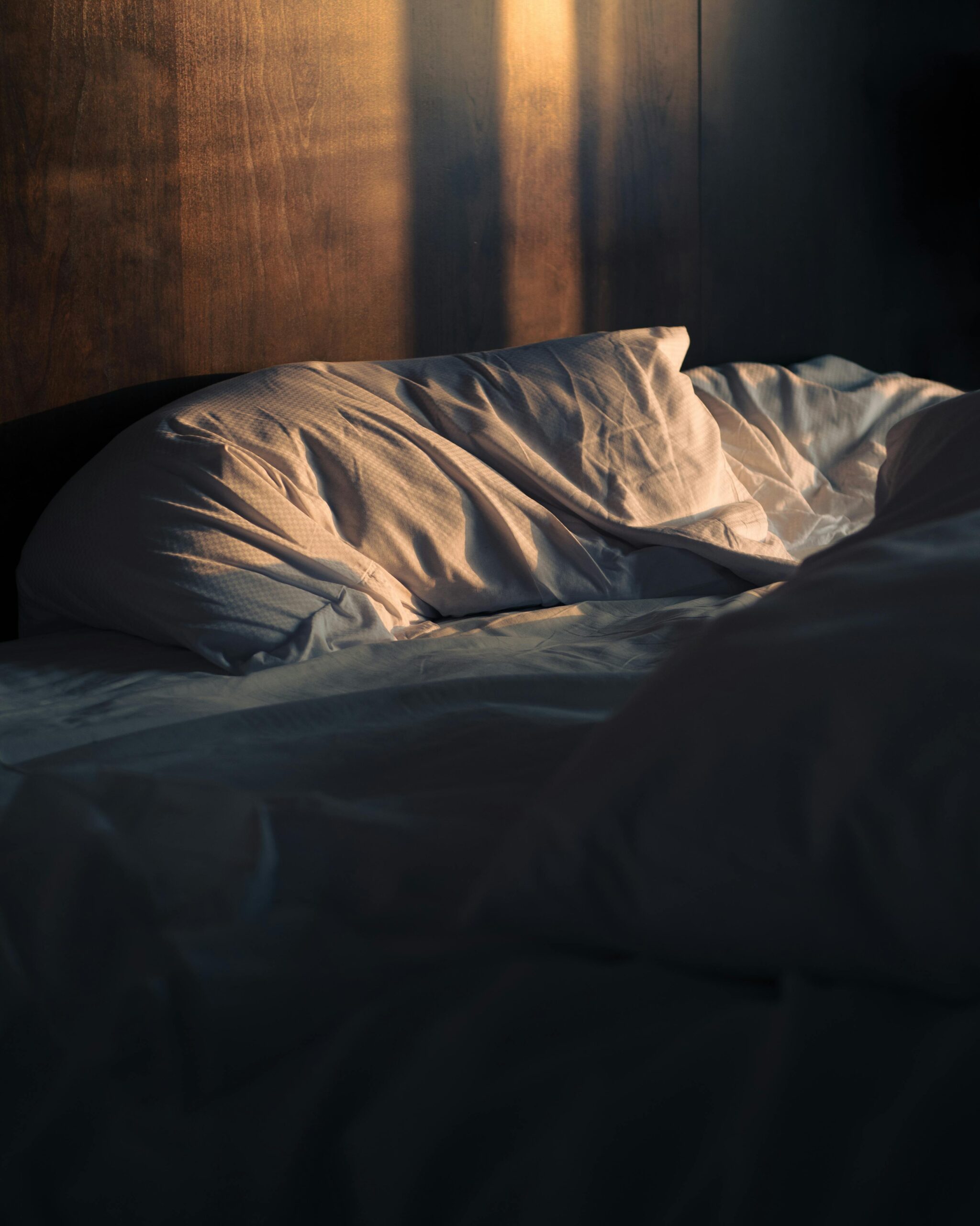 Blick auf ein Bett in einem abgedunkelten Schlafzimmer. Das Kissen sieht etwas platt gedrückt, das Laken verrutscht aus. Die Sonne scheint in das Zimmer herein, an der Rückwand sieht man helle Stellen und Schatten.