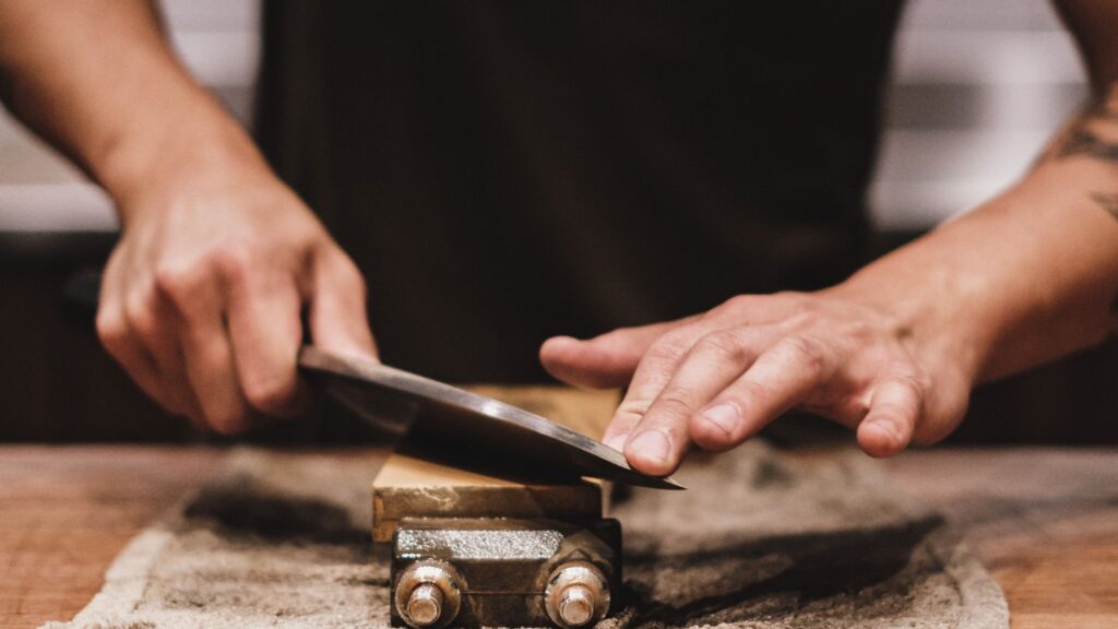 Ein Messer liegt, geführt von einer Männerhand, auf einem eingespannten Holzblock. Unter der Klemme liegt ein etwas schmuddeliger Lappen auf dem Holztisch. Der Mann stabilisiert das Messer mit der linken Hand bei seiner Schnittführung. Er trägt ein schwarzes T-Shirt.