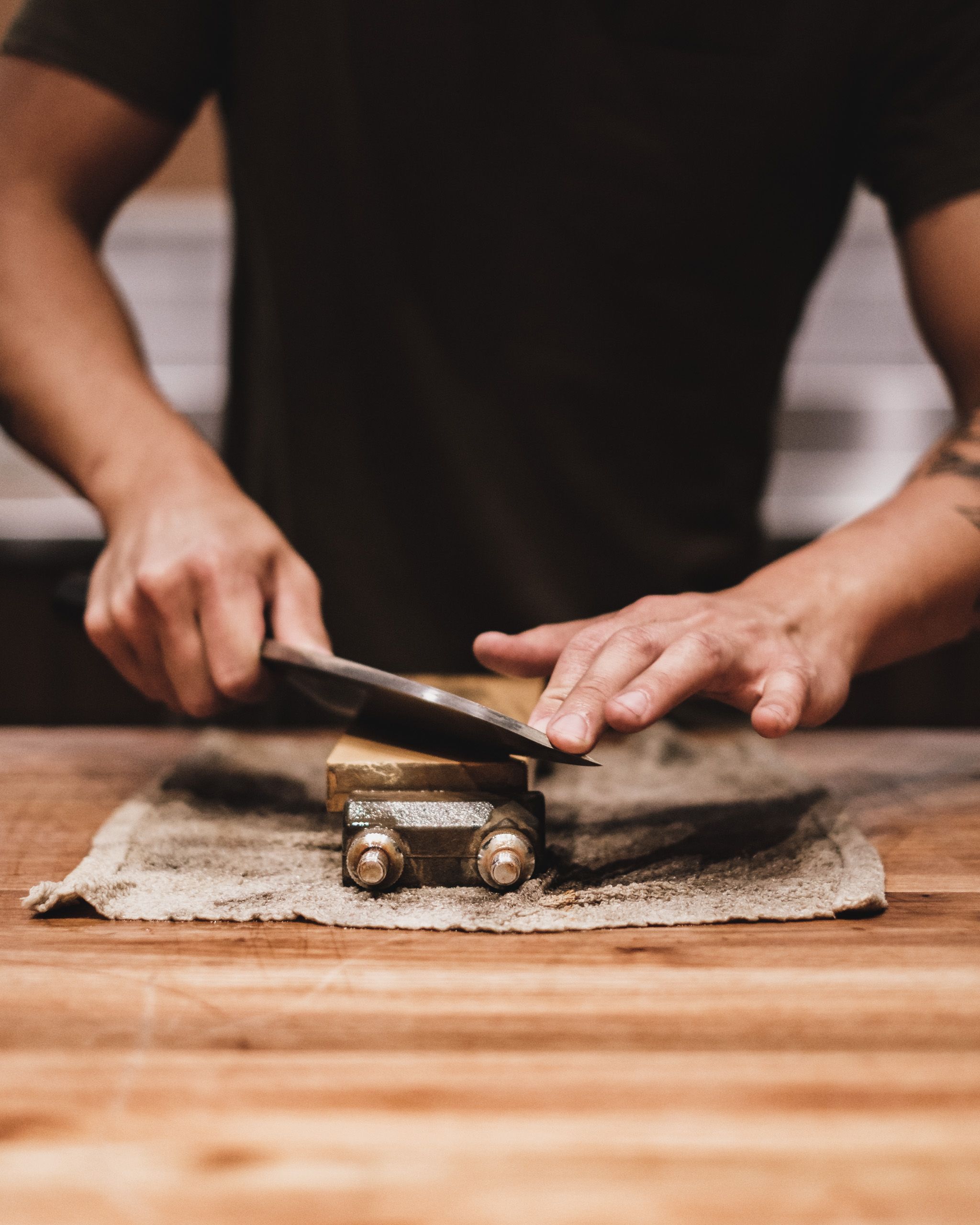Ein Messer liegt, geführt von einer Männerhand, auf einem eingespannten Holzblock. Unter der Klemme liegt ein etwas schmuddeliger Lappen auf dem Holztisch. Der Mann stabilisiert das Messer mit der linken Hand bei seiner Schnittführung. Er trägt ein schwarzes T-Shirt.