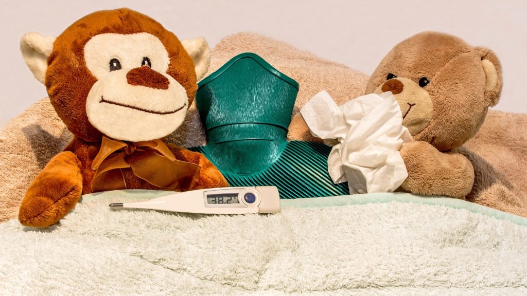 Zwei erkältete Teddybären ruhen sich mit einer Wärmflasche im Bettchen aus, Das Fieberthermometer zeigt 38,2°C an, ein Bärchen schneuzt sich die Nase.