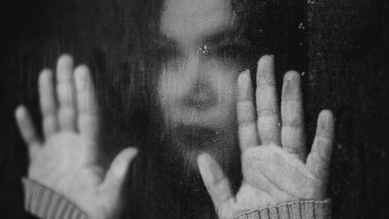 Symbolbild ME/CFS: Frau schaut durch eine verzerrende Fensterscheibe und legt die Handflächen in Höhe ihres Kopfes von innen auf die Scheibe. Sie guckt antriebslos-niedergeschlagen.