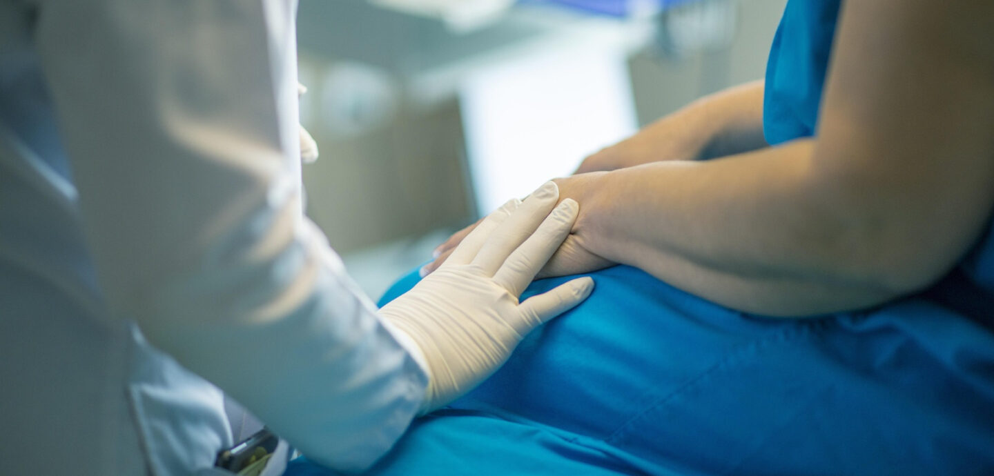 Szene aus dem Krankenhaus. Eine Person in weißem Kittel mit Latexhandschuhen legt seine Hand auf Hand und Oberschenkel einer vermutlich auf einer Liege sitzenden Frau in blauer Kleidung. Der Hintergrund ist unscharf, dürfte aber ein Behandlungszimmer darstellen.