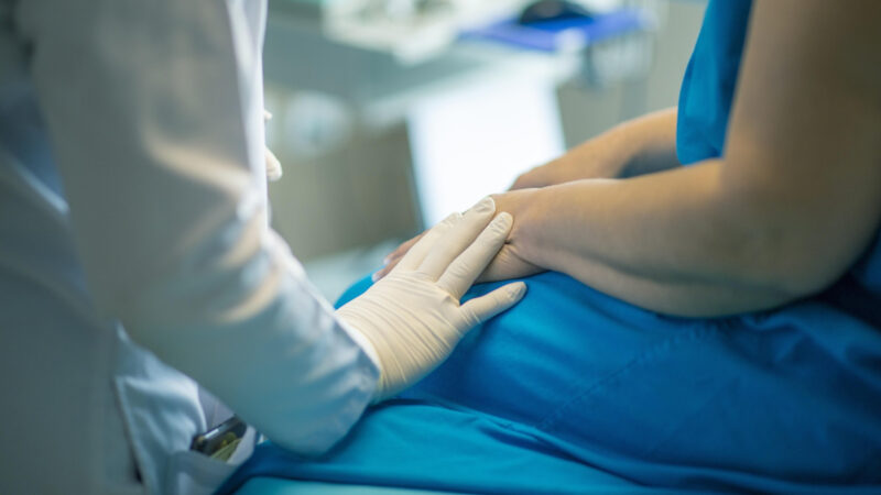 Szene aus dem Krankenhaus. Eine Person in weißem Kittel mit Latexhandschuhen legt seine Hand auf Hand und Oberschenkel einer vermutlich auf einer Liege sitzenden Frau in blauer Kleidung. Der Hintergrund ist unscharf, dürfte aber ein Behandlungszimmer darstellen.